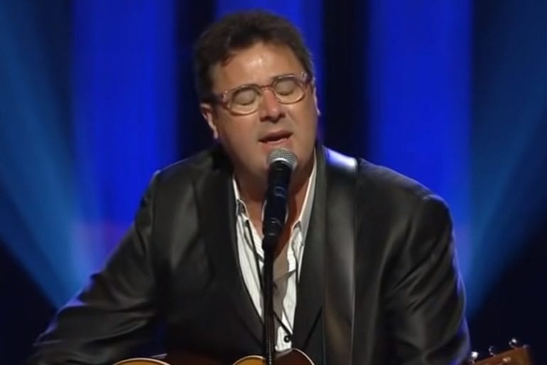 Vince Gill in a suit and glasses speaking into a microphone