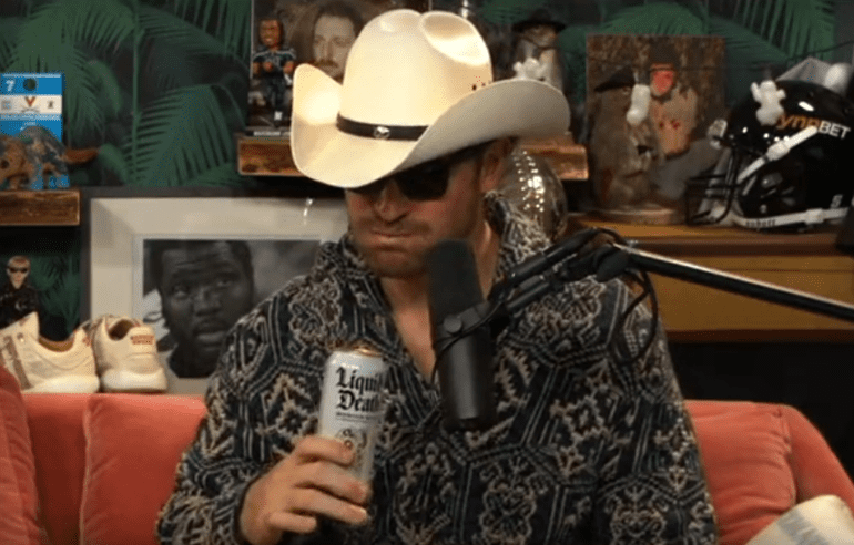 A person wearing a cowboy hat and sunglasses holding a bottle of beer