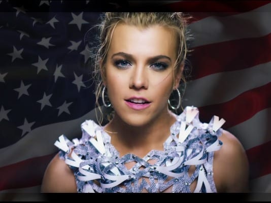 Kimberly Perry with blonde hair in a floral shirt in front of a flag