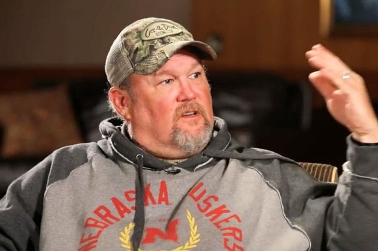 Larry the Cable Guy wearing a hat and holding his hand up