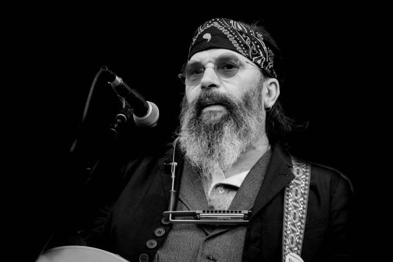 Steve Earle with a beard and a hat holding a microphone