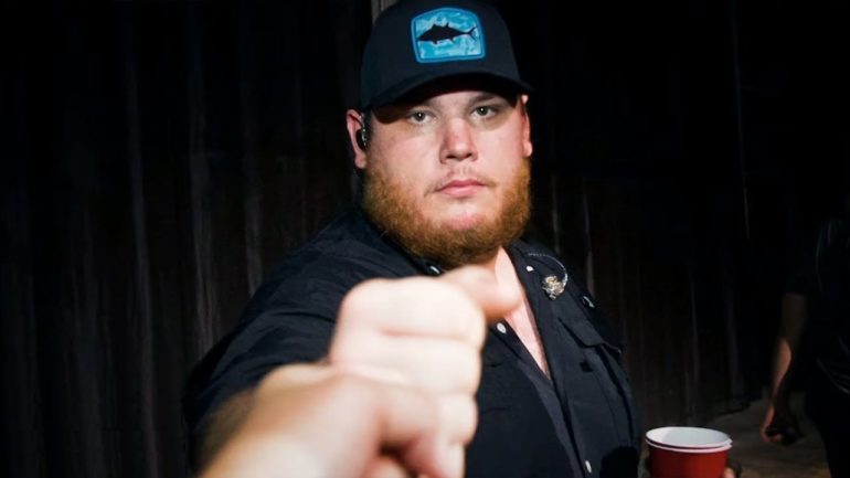 Luke Combs in a hat and a black jacket holding a cup