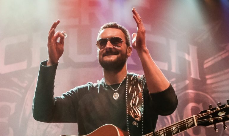 Eric Church with a beard and sunglasses playing a guitar