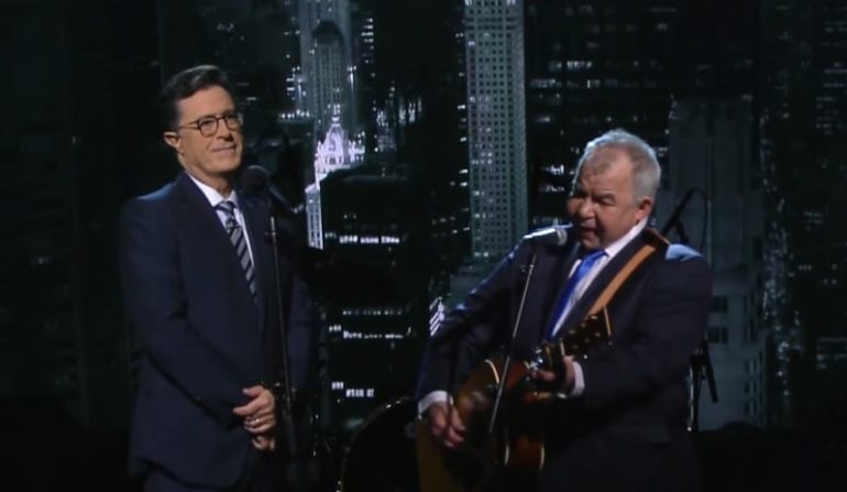 Stephen Colbert speaking into a microphone next to a man in a suit