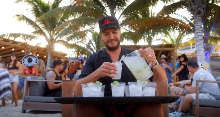 Luke Bryan holding a book and a drink in a crowded outdoor restaurant