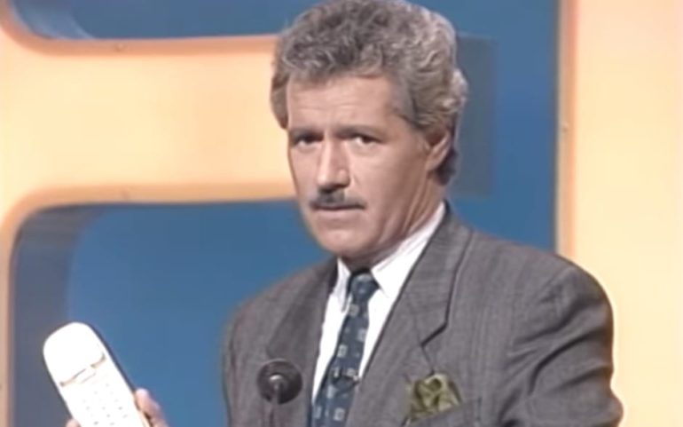 Alex Trebek in a suit and tie holding a cellphone