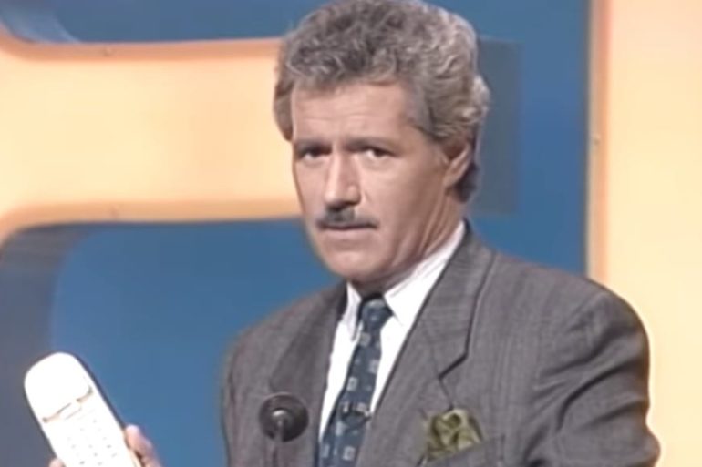 Alex Trebek in a suit and tie holding a cellphone