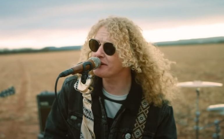 A person with long hair and sunglasses singing into a microphone