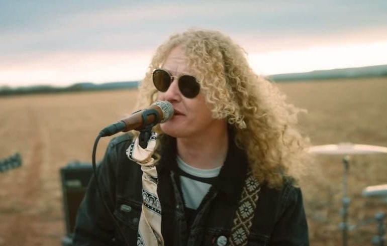 A person with long hair and sunglasses singing into a microphone