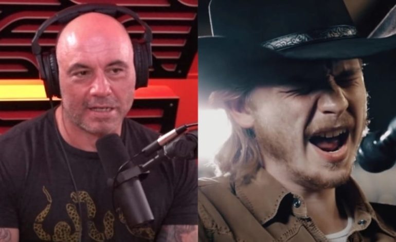 Joe Rogan with a mustache and a hat with a man in a black jacket and a microphone in