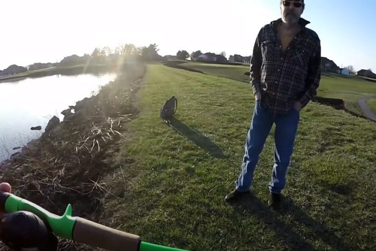 A man holding a gun and standing on grass by a body of water