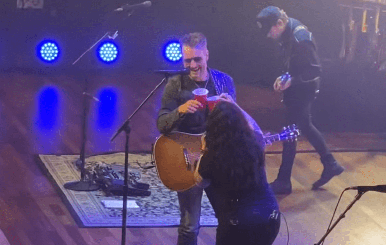 A man singing into a microphone on a stage with a woman holding a microphone and a guitar and