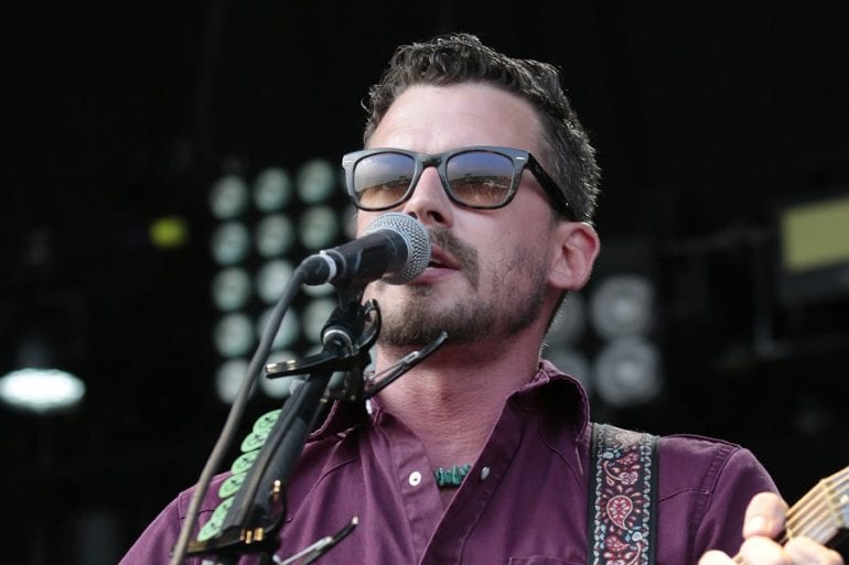A man wearing sunglasses and a purple shirt singing into a microphone