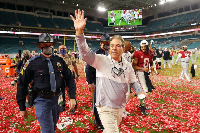 Nick Saban in a suit and tie walking on a field with a crowd of people in the background