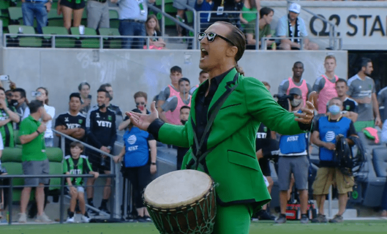 A person wearing a green jacket and sunglasses with a drum in front of a crowd