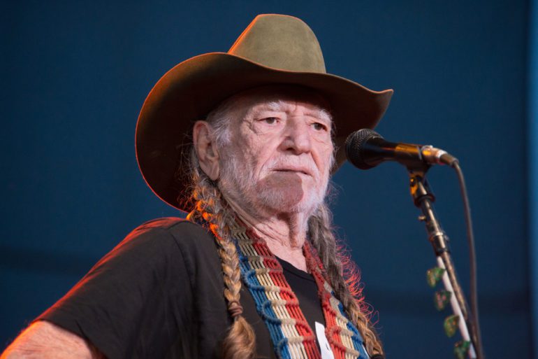 Willie Nelson with a hat and a beard holding a microphone